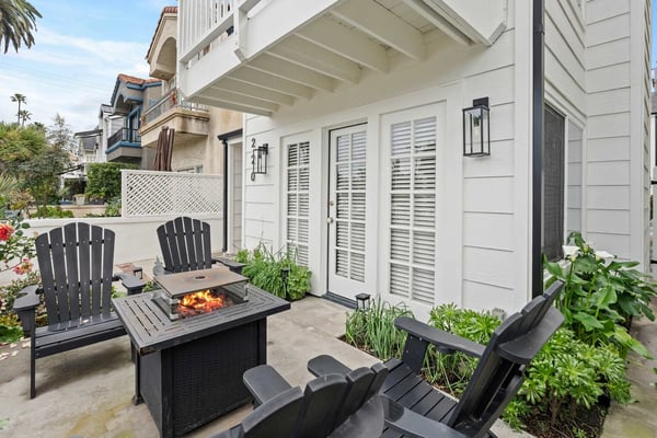 Patio with black Adirondack chairs, fire pit table, potted plants, and white modern house with shuttered doors