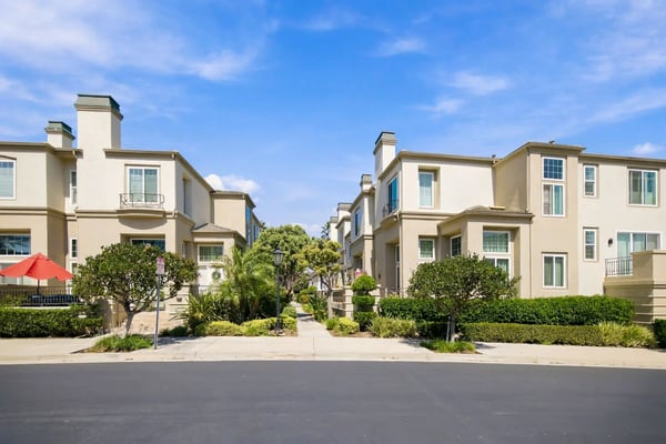 Row of beige townhouses with chimneys, manicured hedges, and palm trees lining a quiet street under blue sky