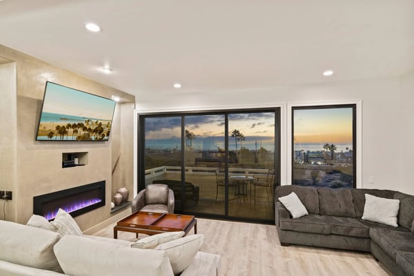 Modern living room with fireplace, ocean and palm tree views through floor-to-ceiling windows, and coastal sunset visible from balcony