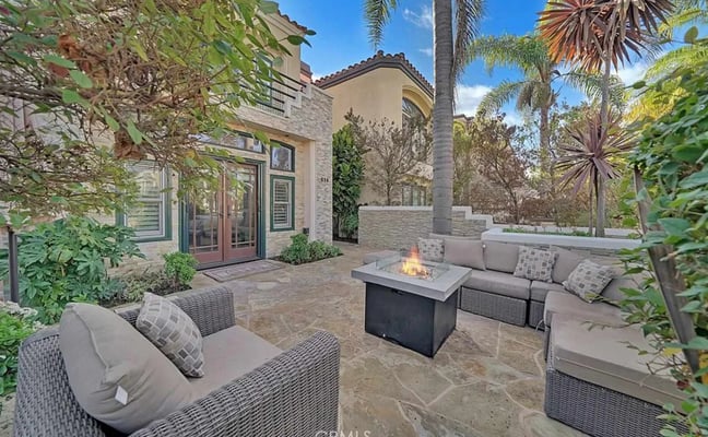 Patio with stone flooring, fire pit table, gray upholstered seating, palm trees, and modern house with arched entry